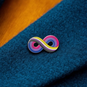 Puede incluir: Un pin de solapa de metal con forma de símbolo de infinito, que presenta un patrón de rayas multicolor con bandas amarillas, blancas, grises, negras, rosas, moradas y azules. El pin está sujeto a una superficie de tela azul verdoso oscuro.