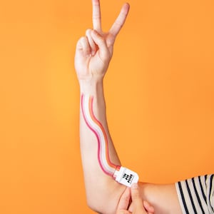 Puede incluir: Un tubo aplicador cosm&eacute;tico blanco con la etiqueta THE PIN PRICK se sostiene contra un antebrazo, creando una franja ondulada y multicolor de pigmento naranja, blanco y rosa que se extiende verticalmente por la piel hacia una mano en se&ntilde;al de paz.