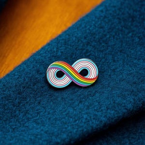 Pode incluir: Um broche de esmalte metálico em forma de símbolo de infinito. O design apresenta um gradiente de arco-íris no lado esquerdo e um padrão de listras em azul claro, rosa e branco no lado direito, tudo contornado por finas bordas de metal prateado.