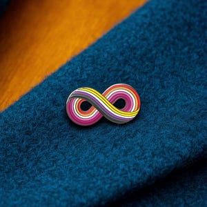 Puede incluir: Un pin de solapa de metal con forma de símbolo de infinito, que presenta un patrón de rayas multicolor en tonos gris oscuro, amarillo, naranja y rosa. El pin tiene un acabado liso y pulido sobre una tela azul oscuro con textura.