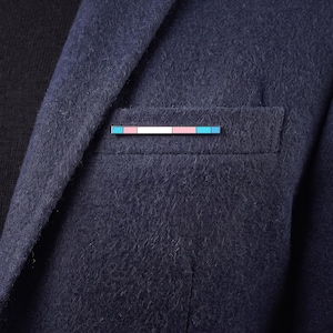 May include: A rectangular lapel pin featuring a horizontal striped pattern in light blue, pink, and white, attached to the breast pocket of a dark navy blue textured wool blazer worn over a black shirt. The pin has a clean, linear design.