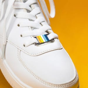 May include: A white sneaker featuring a rectangular metal lace charm threaded onto the white laces. The charm displays a vertical striped pattern in yellow, white, light blue, and dark blue colors, secured by a silver-toned metal frame.