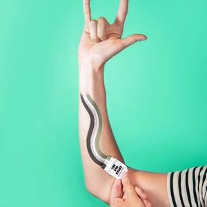 Puede incluir: Un peque&ntilde;o aplicador cosm&eacute;tico blanco con la etiqueta THE PIN PRICK y tapa negra, que traza una l&iacute;nea ondulada de pigmentos verde, oliva y gris oscuro sobre un brazo. La mano est&aacute; posicionada haciendo un gesto de rock and roll.