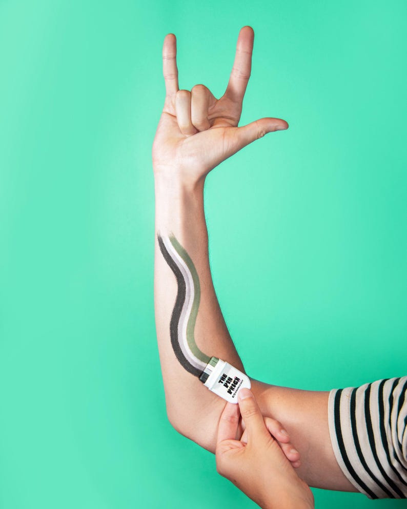 Puede incluir: Un peque&ntilde;o aplicador cosm&eacute;tico blanco con la etiqueta THE PIN PRICK y tapa negra, que traza una l&iacute;nea ondulada de pigmentos verde, oliva y gris oscuro sobre un brazo. La mano est&aacute; posicionada haciendo un gesto de rock and roll.