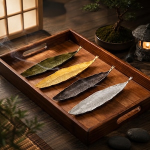 Peut inclure: Un plateau rectangulaire en bois contenant quatre porte-encens allongés en forme de feuille avec des finitions métalliques verte, dorée, bronze foncé et argentée. Chaque support présente une surface texturée avec des nervures visibles.
