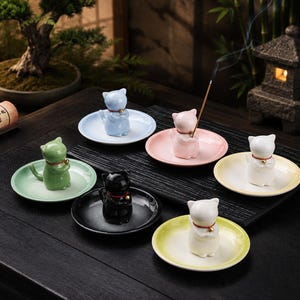 Peut inclure: Six porte-encens en céramique, chacun doté d'une petite figurine de chat avec un collier rouge et une clochette dorée, centrée sur une coupelle ronde assortie. Les chats sont bleu clair, rose, vert, noir, blanc et jaune, avec un bâtonnet.