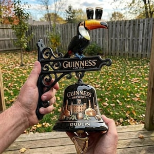 May include: A black metal wall bracket featuring a toucan with two glasses on its beak, the text GUINNESS ESTD 1759, and a hanging black bell with the text GUINNESS BREWERY DUBLIN, a brewery illustration, and a gold-coloured harp clapper.