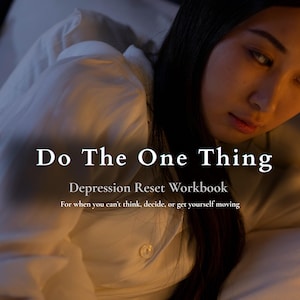May include: A book cover titled Do The One Thing Depression Reset Workbook For when you can't think, decide, or get yourself moving. The cover features a person with long dark hair wearing a white button-down shirt, resting on white bedding.