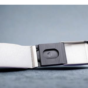 May include: A white fabric belt featuring a rectangular metallic buckle mechanism. The buckle has a dark gray textured central component with a small oval cutout and a silver-toned frame. The belt strap is made of a woven white material.