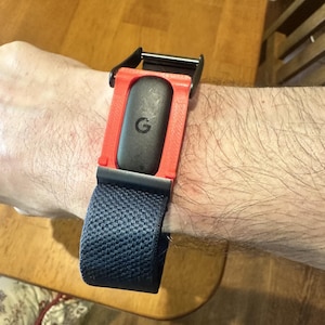 May include: A black fitness tracker with a prominent letter G logo, secured in a bright red 3D-printed plastic housing. The device is attached to a dark blue woven fabric strap with a black metal buckle, worn around a wrist.