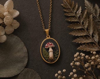 Puede incluir: Un collar con colgante ovalado que presenta un relieve detallado de un hongo rojo y blanco con hojas de helecho verdes sobre un fondo negro, enmarcado en metal dorado y suspendido de una cadena de eslabones dorada a juego.