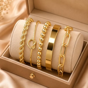 May include: A set of five gold-toned bracelets displayed on a beige velvet jewelry roll. The collection includes a twisted rope chain, a thin bangle with a knot detail, a beaded bracelet, a smooth flat cuff, and a chunky rectangular link chain.