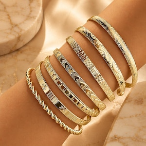 May include: A set of seven gold-toned metal bangles featuring various textures and patterns, including a twisted rope design, star motifs, geometric shapes, and heart cutouts, arranged in a stack on a wrist with a polished metallic finish.