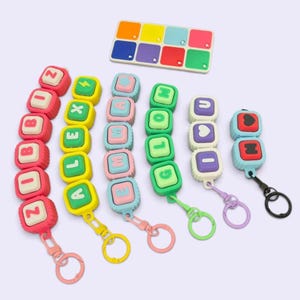 May include: A collection of colourful silicone keychains featuring square blocks with letters, symbols, and hearts, alongside a rectangular silicone tray with eight square compartments. The keychains display various words including ZOOBOZ, ALEX, EMMA, GLOW, and I LOVE U.