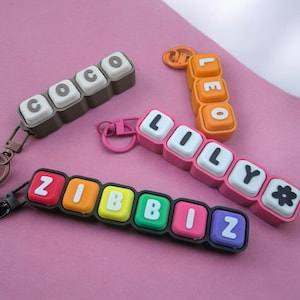 May include: Four custom keychains featuring block-style letters. One brown keychain spells COCO, one orange spells LEO, one pink spells LILY with a flower icon, and one black spells ZIBBIZ in red, orange, yellow, green, pink, and purple blocks.