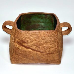 Op de afbeelding: Een vierkante keramische kom met twee handvatten aan de zijkanten, voorzien van een getextureerde, metallic bronskleurige buitenkant en een contrasterende gladde, levendig groene binnenglazuur. Het kleioppervlak oogt ruw en organisch.
