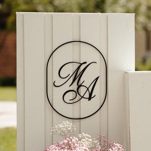 Puede incluir: Cartel de boda blanco con el texto "WELCOME TO THE WEDDING of ARYA & MEAGAN". Un segundo cartel muestra las iniciales "M A" en un óvalo negro. Un jarrón de flores rosas se encuentra en la base de los carteles.