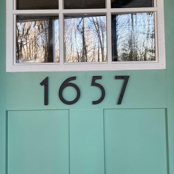 Può includere: Una porta verde menta chiaro con una finestra rettangolare a sei riquadri divisi da cornici bianche. Il numero 1657 è visualizzato in grandi cifre metalliche nere senza grazie, montate orizzontalmente al centro del pannello della porta.