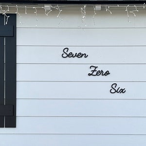 May include: Black cursive metal lettering spelling out the words Seven Zero Six mounted on the white horizontal siding of a house exterior. The text is arranged in three staggered lines between a window with black shutters and a front porch.