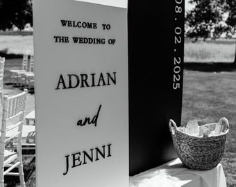 Könnte beinhalten: Ein schwarz-weißes Hochzeits-Willkommensschild mit dem Text Welcome to the wedding of Adrian and Jenni und dem Datum 08. 02. 2025. Das Schild besteht aus einer hohen weißen rechteckigen Platte neben einer höheren schwarzen rechteckigen Platte.