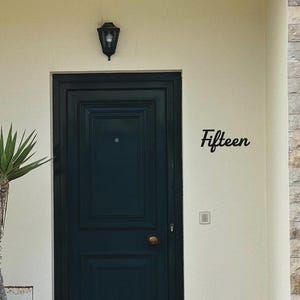 May include: A dark green paneled door with a round brass knob and a black wall-mounted light fixture above. To the right, the word Fifteen is written in black script. A tall green plant in a terracotta pot sits to the left of the door.