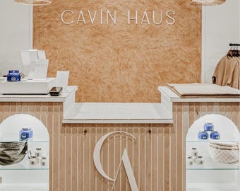 Könnte beinhalten: Ein Verkaufstresen mit einer Basis aus hellen Holzlatten und einem weißen CA-Logo, einer strukturierten beigen Wand mit dem weißen Schriftzug CAVIN HAUS, zwei Glaspendelleuchten und zwei gewölbten Nischen mit kleinen blauen Gläsern und Taschen.
