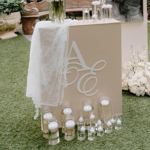 May include: An outdoor wedding reception setup featuring a beige pedestal with white floral arrangements, candles in glass holders, and a large black and white photo. The pedestal has the initials "A" and "C" in a cursive font.