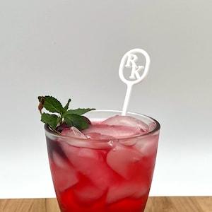 Puede incluir: Un cóctel rojo refrescante en un vaso transparente con cubitos de hielo y una ramita de menta. Un agitador blanco con las letras "R" y "K" está en la bebida. La bebida está sobre una superficie de madera.