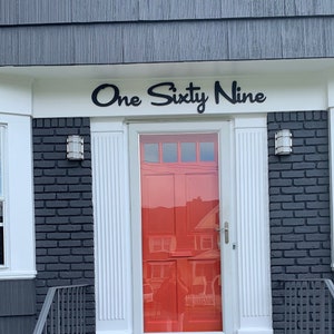 May include: A red front door with a white frame and a black doormat. The door is on a house with black brick and white trim. The house number is "One Sixty Nine" in black letters above the door.