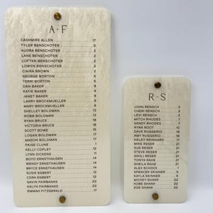 May include: Two rectangular off-white acrylic signs with a marbled texture. The larger sign is labeled A-F and the smaller sign is labeled R-S. Both feature lists of names followed by numbers, secured with small gold-toned circular metal fasteners.
