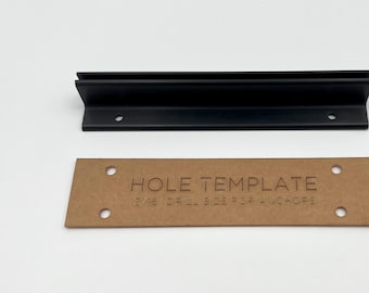 Może przedstawiać: Czarny metalowy wspornik półki z szablonem otworów i osprzętem montażowym. Szablon w kolorze brązowym z napisem "HOLE TEMPLATE" i "3/16 DRILL SIZE FOR ANCHORS". Zawiera śruby i kołki.