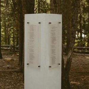 May include: Two tall, white, rectangular pillars stand upright. The left pillar features the black text "A LOVE SET IN STONE" vertically. The right pillar displays two transparent panels with printed text attached by black hardware. A floral arrangement sits at the base.