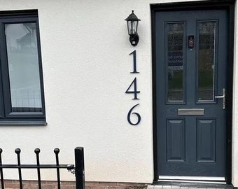 Puede incluir: Una casa blanca con una puerta negra y los números negros 146 en el lateral. La puerta tiene un panel de vidrio con una manija de puerta plateada. Hay una lámpara negra sobre la puerta.