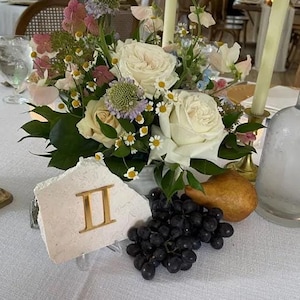 May include: A white table setting with a floral centerpiece, a white ceramic number "II" table number, a bottle of water, grapes, and a pear.