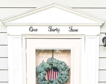 May include: A light pink door with a wreath and an American flag. The wreath is made of green leaves and the flag is visible through the glass. The door is framed by white trim and the words "One Forty Five" are above the door.