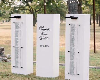 Könnte beinhalten: Drei weiße, rechteckige Hochzeits-Sitzplan-Displays mit Gästenamen. Das mittlere Display hat den Text "Happily Ever Walter" und das Datum "10.12.2024". Die Displays befinden sich im Freien auf einer Rasenfläche, mit Lichterketten und Bäumen im Hintergrund.
