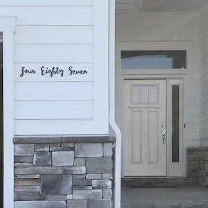 May include: A white house with a gray stone base and a white door with a glass window above it. The house number "Four Eighty Seven" is displayed on the white siding.