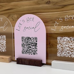 Op de afbeelding: Drie acrylplaten met afgeronde toppen, elk met de tekst "Let's get social" in witte schrijfletters. De platen hebben een QR-code erop gedrukt en staan op houten standaardjes. De platen zijn roze, helder en mat.