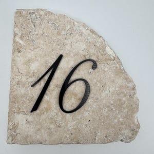 Può includere: Una targa con numero civico che presenta il numero 16 nero in un carattere corsivo, montata su un pezzo di pietra travertino beige naturale dai bordi grezzi, con una superficie testurizzata e porosa e una forma semicircolare irregolare.