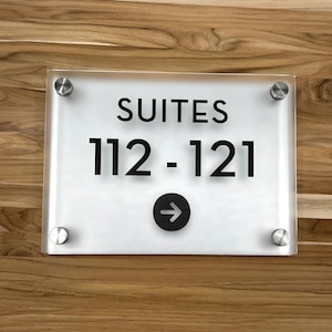 Op de afbeelding: Wit acryl bord met zwarte tekst en een zwarte pijl. Het bord heeft de tekst "SUITES 112-121" en heeft vier zilveren schroeven.