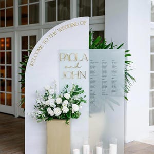 May include: A wedding reception display with the text "WELCOME TO THE WEDDING OF PAOLA and JOHN" in gold lettering. The display includes a floral arrangement of white flowers and candles.
