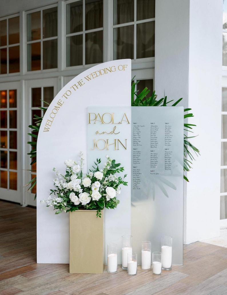May include: A wedding reception display with the text "WELCOME TO THE WEDDING OF PAOLA and JOHN" in gold lettering. The display includes a floral arrangement of white flowers and candles.