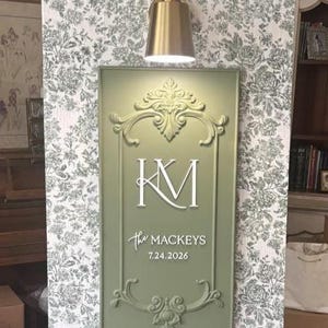 May include: A rectangular sage green sign with a raised ornate border, featuring the white monogram KM, the text The MACKEYS, and the date 7.24.2026. The sign is mounted on a white wall with a green floral pattern and topped with a gold wall sconce.