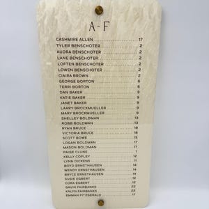 May include: A rectangular off-white acrylic sign with a marbled texture featuring the text A-F at the top, followed by a list of names and corresponding numbers. The sign is secured with two gold-toned circular metal fasteners at the top and bottom.