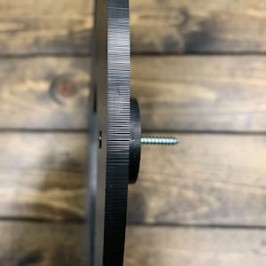 May include: Black metal disc with a textured edge and a screw in the center. The disc is mounted on a wooden surface.