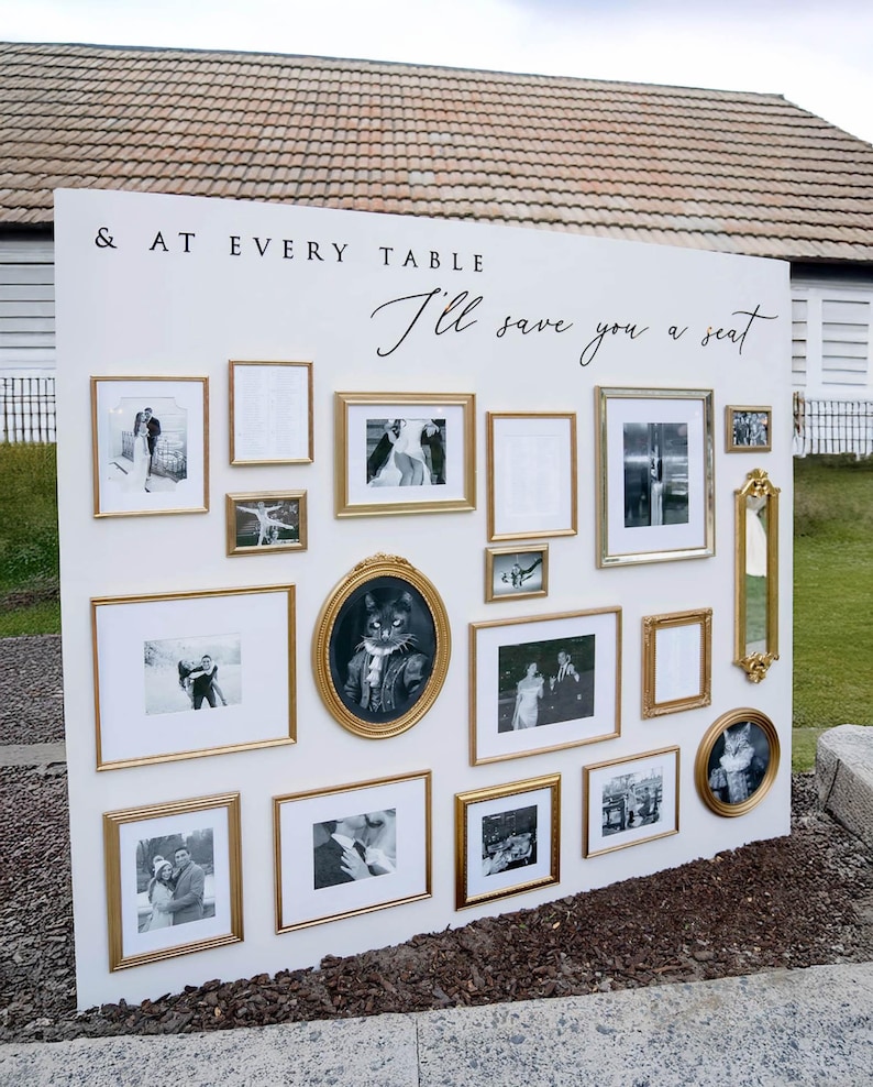 Puede incluir: Un gran panel expositor blanco con el texto & AT EVERY TABLE I'll save you a seat, adornado con un surtido de marcos dorados de diversas formas y tama&ntilde;os, algunos de los cuales contienen fotograf&iacute;as y retratos en blanco y negro.