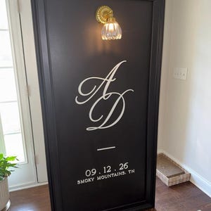 May include: A tall, rectangular black sign with a thick frame, featuring a gold-toned wall sconce with a ribbed glass shade at the top. The sign displays the white monogram letters AD, the date 09.12.26, and the text SMOKY MOUNTAINS, TN.