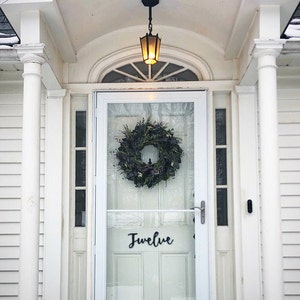May include: A white front door with a glass storm door. The storm door has a wreath made of greenery and a sign that says "Twelve".