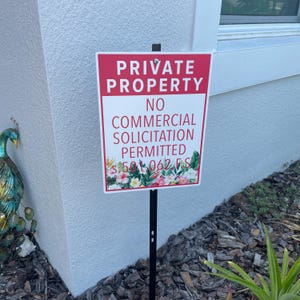May include: A rectangular metal sign on a black stake featuring a red header with the white text PRIVATE PROPERTY and a white lower section with the red text NO COMMERCIAL SOLICITATION PERMITTED s 501.062 FS above a colorful floral pattern.