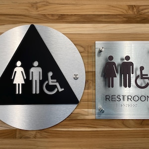 Peut inclure: Deux panneaux en métal argenté avec des symboles noirs pour des toilettes. Le panneau de gauche est rond avec un triangle noir à l'intérieur. Le triangle contient des symboles pour une femme, un homme et une personne en fauteuil roulant. Le panneau de droite est rectangulaire avec le mot "RESTROOM" en lettres noires et le braille en dessous.